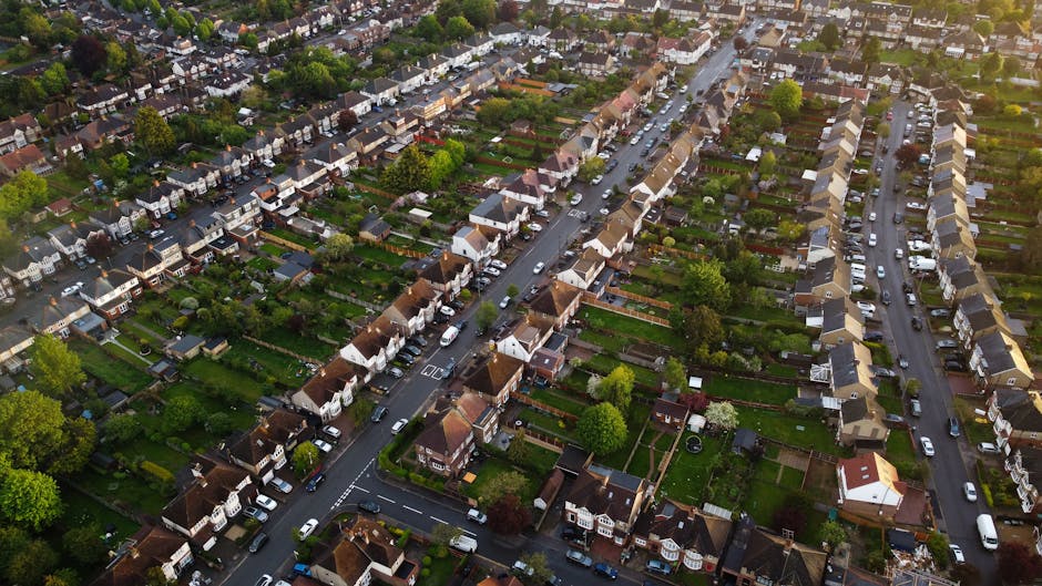 An aerial view of a residential neighbourhood showing narrow lanes lined with neatly arranged terraced houses, detached and semi-detached homes with pitched roofs, and well-maintained gardens. Cars are parked along the streets and driveways, and the scene includes lush green trees and lawns. The environment appears to be a quiet suburban area, with a focus on the layout of streets and housing. This setting illustrates typical elements involved in house removals and home relocation processes, such as the layout of properties and access routes for moving vehicles, with a van possibly visible nearby. The image supports content related to furniture transport, packing, and logistical planning for estate moves as discussed on the Aldersbrook Estate moves page, with natural integration for a company like Man with Van Wanstead specializing in removals.