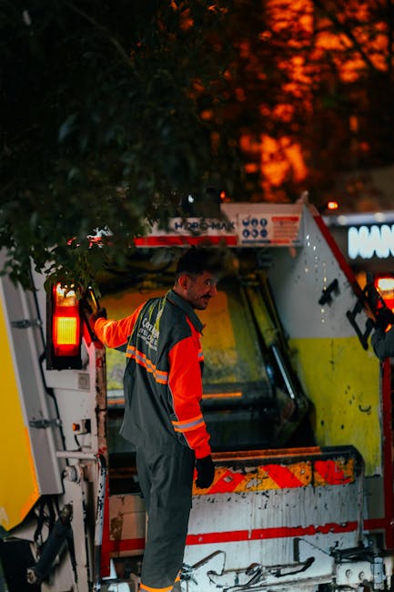 A male worker wearing high-visibility orange and grey work clothing is engaged in a home relocation task outdoors during evening or night, with the sky illuminated by orange sunset or streetlights. He is standing next to a large, white and yellow garbage or recycling truck, which is parked on a paved area near a building. The worker appears to be either inspecting the vehicle or preparing for the loading process, with his right hand resting on the side of the truck. The surrounding environment includes a tree partially obscuring the view and ambient lighting from the vehicle's tail lights and nearby sources. This scene exemplifies the logistics involved in professional removals and waste management, supporting services such as house removals and clearance often handled by companies like Man with Van Wanstead, which specialise in furniture transport, packing, and loading operations.
