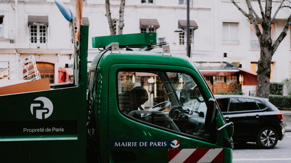 Front-side view of a green removal truck parked on a street in Wanstead, with the driver visible through the side window. The truck features branding including the logo of Man with Van Wanstead and the words 'Propreté de Paris' and 'Mairie de Paris.' The vehicle is equipped for furniture transport and home relocation, with a loading area at the rear where items such as cardboard boxes, plastic containers, and wrapped furniture are visible. The boxes are stacked neatly, some wrapped in packing materials like bubble wrap or fabric covers. The truck is positioned close to a residential building with white exterior walls, and nearby trees with bare branches imply the scene is in late autumn or winter. Other parked cars are visible along the street, and the lighting appears natural, indicating daytime with an overcast sky. This image captures the loading process involved in domestic removals and packing tasks performed by professional services like Man with Van Wanstead, highlighting the logistical aspects of home relocation.