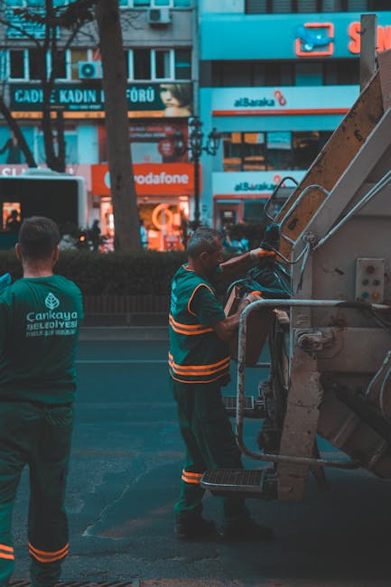 A male worker wearing high-visibility orange and grey work clothing is engaged in a home relocation task outdoors during evening or night, with the sky illuminated by orange sunset or streetlights. He is standing next to a large, white and yellow garbage or recycling truck, which is parked on a paved area near a building. The worker appears to be either inspecting the vehicle or preparing for the loading process, with his right hand resting on the side of the truck. The surrounding environment includes a tree partially obscuring the view and ambient lighting from the vehicle's tail lights and nearby sources. This scene exemplifies the logistics involved in professional removals and waste management, supporting services such as house removals and clearance often handled by companies like Man with Van Wanstead, which specialise in furniture transport, packing, and loading operations.
