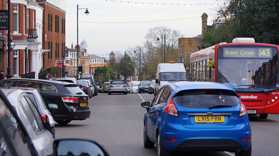 A street scene in Wanstead during daytime showing multiple parked vehicles along both sides of the road, including a prominent blue hatchback with a yellow registration plate 'LV15 FRN' parked on the right side of the street. A red Metroline bus with route number 143 and destination 'Brent Cross' is traveling along the road, passing the parked cars. Behind the bus, a white van is visible, positioned next to the pavement near residential buildings with brick facades and white window frames. Street lamps and overhead electrical wires run across the street, with leafless trees in the background suggesting late autumn or early winter. The scene highlights typical urban parking and moving logistics considerations, relevant to house relocations and furniture transport in Wanstead, as addressed by Man with Van Wanstead’s removals services.