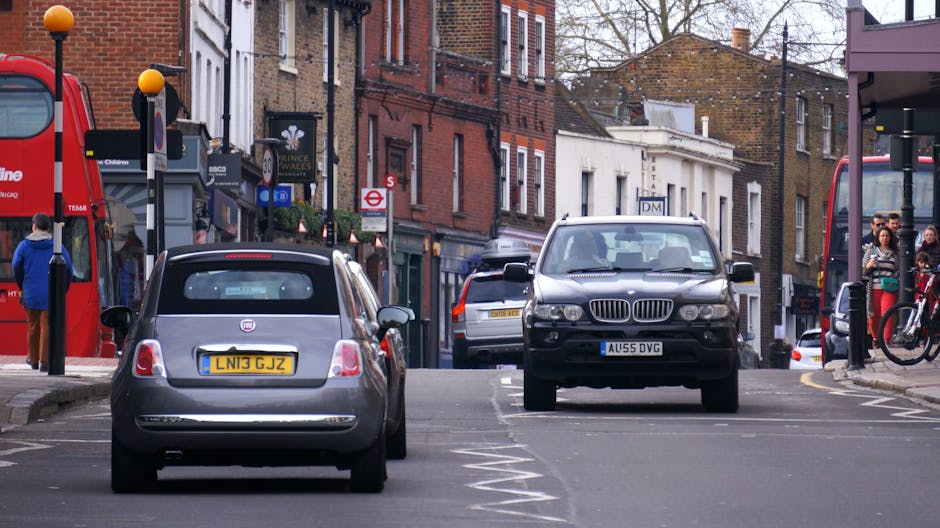 A residential street in Wanstead with parked cars lining both sides of the paved road, including a variety of vehicles such as small hatchbacks, vans, and larger commercial vans, some with open rear doors revealing cardboard boxes, plastic wrapping, and packed furniture ready for home relocation. The scene shows an overcast sky with gray clouds, and one side of the street features terraced houses with red brick facades, small front gardens, and trees, some with vibrant autumn foliage. Utility poles and overhead power lines extend along the street, with a large, leafy green tree at the end of the street. The image captures the busy environment typical of moving logistics, where careful planning by companies like Man with Van Wanstead ensures smooth furniture transport and loading processes during a house move, with attention to parking regulations to avoid fines during relocation.