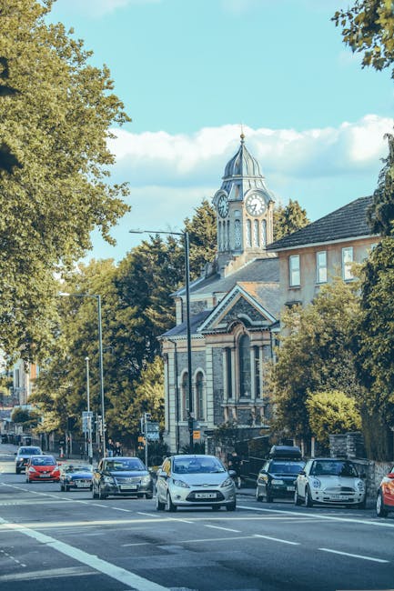 A street scene in Wanstead showing parked cars along the curb, with a large historic church featuring a clock tower and a spire in the background, surrounded by leafy trees under a blue sky with some clouds. The image captures the exterior environment where house removals and moving services by Man with Van Wanstead are often conducted, highlighting urban logistics and transport elements like vehicle parking, street lighting, and building facades relevant to home relocation and furniture transport processes in the Wanstead area.