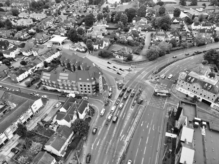 A street scene in Wanstead showing parked cars along the curb, with a large historic church featuring a clock tower and a spire in the background, surrounded by leafy trees under a blue sky with some clouds. The image captures the exterior environment where house removals and moving services by Man with Van Wanstead are often conducted, highlighting urban logistics and transport elements like vehicle parking, street lighting, and building facades relevant to home relocation and furniture transport processes in the Wanstead area.