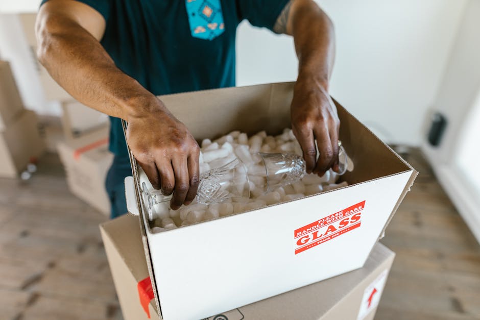 A person wearing a blue shirt is placing a clear glass bottle into a cardboard box filled with white packing peanuts on a wooden floor indoors. The box is labeled with red stickers that indicate it contains fragile glass items, emphasizing careful handling during home relocation or furniture transport. In the background, there are additional boxes and packing materials, suggesting a packing or loading process as part of a house move. The lighting is natural, highlighting the details of the packing materials and the individual's hands, which are carefully inserting the glass bottle to prevent breakage during the moving and delivery process. Man with Van Wanstead specializes in removals, including packing and moving delicate items like antiques and glassware, ensuring secure transportation during home relocations.
