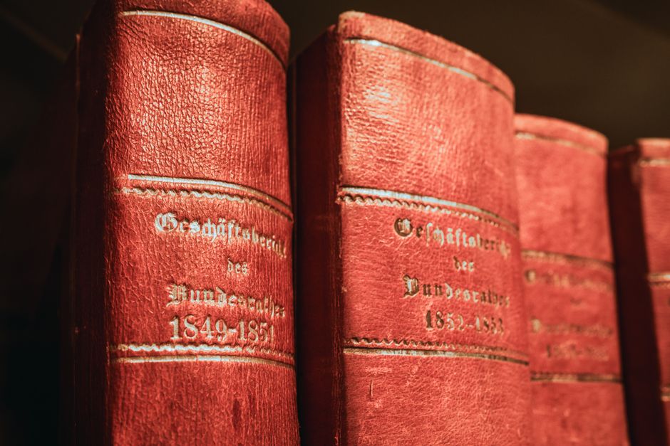 A close-up view of several antique red hardcover books with embossed silver titles and decorative borders, stacked vertically on a dark wooden shelf. The books are well-preserved, showing textured leather covers and gold-embossed German text, indicating they are historical volumes related to 19th-century German history, including dates from 1849 to 1851. The lighting highlights the rich color and intricate details of the book covers. In the background, a portion of a room interior is visible, possibly a storage or study area used for house removals. The scene captures the careful handling and packing process involved in home relocation, especially when moving valuable or delicate items such as antique books, which [COMPANY_NAME] may assist with during furniture transport or packing and moving services. The environment appears organized, with the focus on securely loading or unpacking these items for a smooth transition, reflecting expertise in delicate relocation tasks as outlined in the context of legal tips for moving antiques in Wanstead, WANSTEAD.