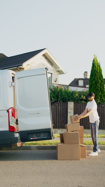 A man wearing a white t-shirt and glasses is outdoors, standing beside a white moving van with its rear door open, as part of a home relocation process. He is placing a cardboard box onto a stack of t