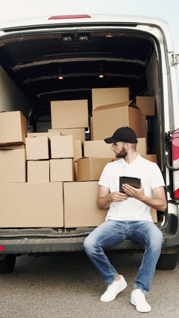 A man wearing a white t-shirt and glasses is outdoors, standing beside a white moving van with its rear door open, as part of a home relocation process. He is placing a cardboard box onto a stack of t