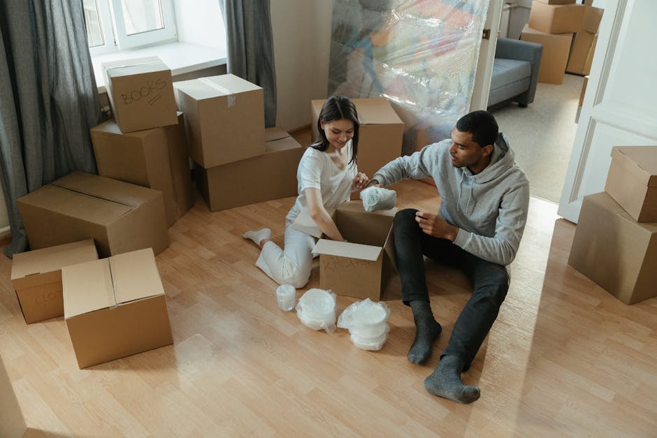 A young woman and a young man are sitting on a light wooden floor inside a room, surrounded by numerous cardboard boxes, some labeled with words like 'books,' and packing materials such as bubble wrap. The woman is holding a piece of bubble wrap, while the man is holding a box with packing paper inside, possibly preparing for a home relocation. The room has a grey sofa near the background, a window with grey curtains, and several cardboard boxes of various sizes stacked against the wall, indicative of a packing and moving process. The space is illuminated by natural light coming through the window, and additional boxes are visible in an adjacent room through an open doorway. This scene showcases the packing stage of furniture transport and house removals, with the individuals engaged in carefully handling packing materials to secure belongings for the move, reflecting typical home relocation logistics supported by Man with Van Wanstead.