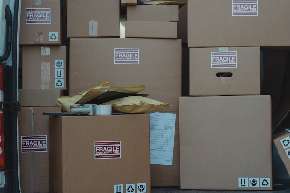 A collection of brown cardboard moving boxes of various sizes, stacked and arranged inside the open rear of a van, ready for home relocation. Several boxes have 'Fragile' labels, and some display handling symbols such as 'this side up,' 'recycle,' and 'protect with care.' There are partially opened padded mailing envelopes on top of a medium-sized box, along with packing tape and a clipboard with a document attached. The boxes are positioned on a flat surface within the van, which is parked on a paved area. The image reflects the careful packing phase of furniture transport and the logistics involved in packing and moving services, as provided by [COMPANY_NAME], to comply with Redbridge Council permits for Wanstead moving vans. The scene highlights the importance of secure loading and organized packaging in the house removals process.