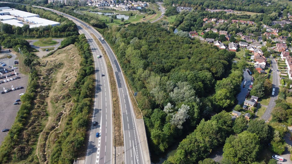 Aerial view of a dual carriageway bordered by dense green woodland on one side and a residential neighbourhood with houses and winding streets on the other, taken during daylight with clear visibility. The road is lined with white dashed lines and several cars are seen traveling along it, with a few parked along the nearby parking lot visible on the left. The woodland area features a mix of deciduous trees with lush foliage, contrasting against the urban surroundings. The residential area includes detached and semi-detached houses with gardens, some with visible driveways, and the scene captures a moment of typical city infrastructure and natural landscape co-existence. This image exemplifies the logistical environment in which house removals and furniture transport services, such as those offered by Man with Van Wanstead, are carried out, highlighting the importance of efficient relocation planning in built-up and green areas during home moving projects.