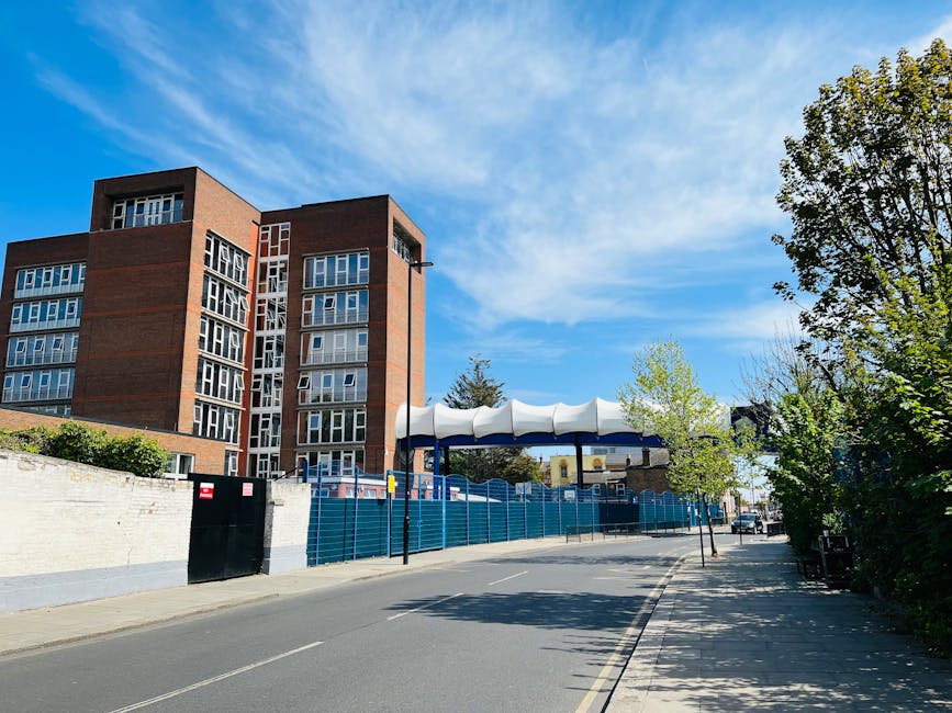 A wide view of a residential street during daytime featuring a multi-storey brick building on the left, with large windows and a modern design. In the foreground, there is a white wall with a black gate, and behind it, a blue construction fence surrounds a site with some equipment partially visible. To the right, several small trees with green foliage line the pavement, casting shadows on the street. Overhead, a blue sky with some wispy clouds is visible. Visible on the right side are some parked vehicles and a pedestrian pathway, suggesting an urban environment suitable for home relocation or furniture transport. The scene captures the environment where Man with Van Wanstead operates, with details indicating ongoing building or renovation work that might impact moving logistics connections, such as suspensions on Redbridge Road for large Wanstead moves.