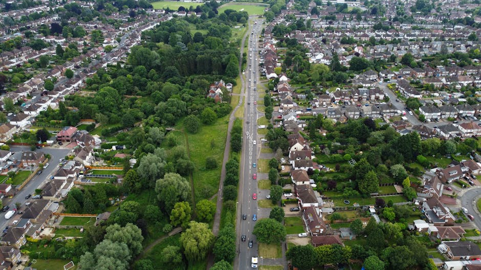 Aerial view of a dual carriageway bordered by dense green woodland on one side and a residential neighbourhood with houses and winding streets on the other, taken during daylight with clear visibility. The road is lined with white dashed lines and several cars are seen traveling along it, with a few parked along the nearby parking lot visible on the left. The woodland area features a mix of deciduous trees with lush foliage, contrasting against the urban surroundings. The residential area includes detached and semi-detached houses with gardens, some with visible driveways, and the scene captures a moment of typical city infrastructure and natural landscape co-existence. This image exemplifies the logistical environment in which house removals and furniture transport services, such as those offered by Man with Van Wanstead, are carried out, highlighting the importance of efficient relocation planning in built-up and green areas during home moving projects.