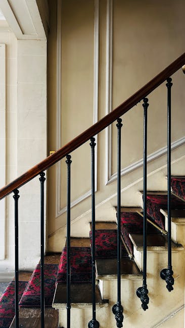 An interior view of a staircase in a Wanstead Victorian home, showing a wooden banister with a polished finish and black metal spindles. The staircase is carpeted with a red and black patterned runner, which extends partially up the steps. The wall beside the staircase features cream-colored paneling with decorative molding, and a decorative wooden skirting board runs along the base. Natural light illuminates the area, highlighting the intricate details of the banister and the texture of the carpet. The image reflects a typical domestic environment where home relocation or furniture transport services, such as those provided by Man with Van Wanstead, might be involved in navigating staircase challenges during a house move.