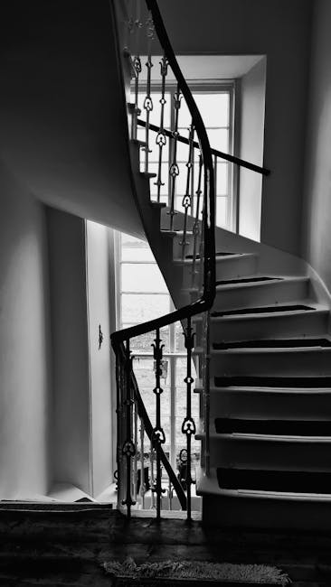 A black-and-white photograph of a winding staircase inside a Wanstead Victorian home, featuring a metal balustrade with decorative spindles and a polished handrail. The staircase curves upward and is positioned next to a large, multi-pane window that allows natural light to illuminate the steps and surrounding area. The stairs are made of wood, with a smooth surface and visible grain, and the treads are supported by stringers along the wall. In the background, the upper landing and part of the interior wall are visible, along with the window's grid of panes. The image captures the architectural details and spatial layout characteristic of historic homes in Wanstead, relevant to home relocation or furniture transport processes. Man with Van Wanstead’s removal services often involve navigating such features when planning moves involving staircases in Victorian properties.