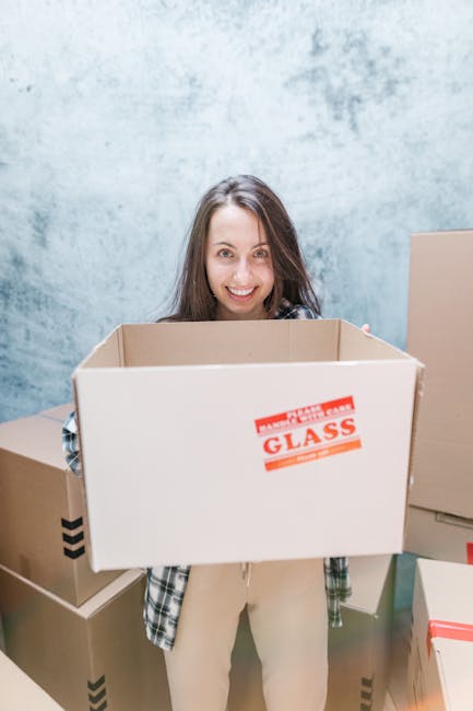 A young woman with long brown hair, smiling and holding an empty cardboard box labeled 'FRAGILE GLASS' in red text, standing indoors against a textured light blue wall. She is dressed casually in a plaid shirt and light-colored trousers. Surrounding her are several other packed boxes of varying sizes, some with black arrow markings indicating orientation, positioned on the floor and nearby surfaces. This scene depicts the packing process associated with home relocation or furniture transport, with the woman preparing items for moving. The environment suggests an organized packing and loading process typical of professional removals services, such as those offered by Man with Van Wanstead, during a house move or furniture transport to or from a property near Wanstead Flats. The lighting is neutral and functional, emphasizing the practical aspects of household packing and moving logistics.