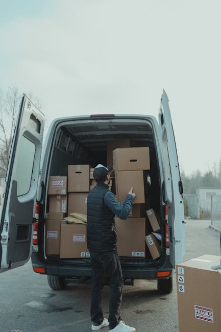 A man stands beside an open van loaded with several cardboard boxes, some of which are taped and stacked. He is wearing a beige knit beanie, a green padded vest over a dark jacket, and dark trousers w